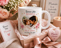 Peut inclure: Une tasse en céramique blanche avec une photo en forme de cœur d'une mère et d'un enfant, entourée de motifs floraux. La tasse porte l'inscription "Bonne Fête Maman" et "Léa & Emma on t'aime très fort". Un coffret cadeau avec un objet en forme de cœur et une carte sont également présents.