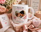 Peut inclure: Une tasse en céramique blanche avec une photo en forme de cœur d'une mère et d'un enfant, entourée de motifs floraux. La tasse porte l'inscription "Bonne Fête Maman" et "Léa & Emma on t'aime très fort". Un coffret cadeau avec un objet en forme de cœur et une carte sont également présents.