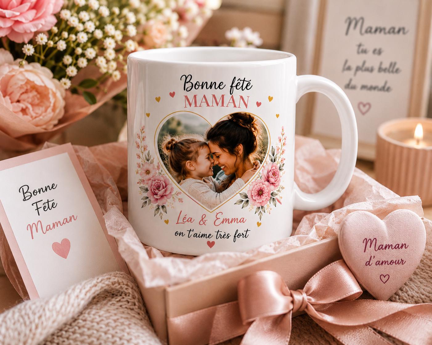 Peut inclure: Une tasse en céramique blanche avec une photo en forme de cœur d'une mère et d'un enfant, entourée de motifs floraux. La tasse porte l'inscription "Bonne Fête Maman" et "Léa & Emma on t'aime très fort". Un coffret cadeau avec un objet en forme de cœur et une carte sont également présents.