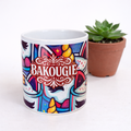 Mug blanc avec illustration multicolore et logo BAKOUGIE au centre, photographie de face sur fond clair avec une plante en pot a l arriere plan