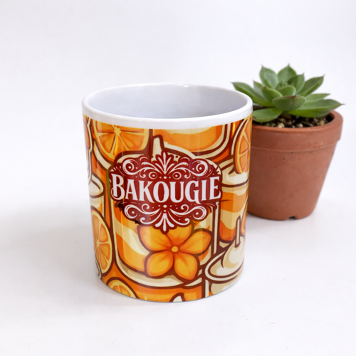 Mug blanc avec decor orange et logo BAKOUGIE au centre, photographie sur fond clair avec une plante grasse en pot a droite