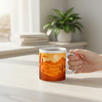 Main tenant un mug blanc avec une illustration de paysage desertique orange avec cactus, avec de la vapeur visible, sur une table claire pres d une fenetre et d une plante