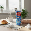 Main tenant un tumbler blanc avec decor bleu BAKOUGIE et motifs de nuages, pose sur une table claire avec un croissant, une tasse fumante et de petits elements blancs decoratifs