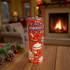 Tumbler blanc avec decor orange BAKOUGIE et paille metallique, pose sur une table en bois dans une ambiance festive avec pommes de pin, branches de sapin et cheminee allumee en arriere-plan