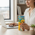 Personne tenant un tumbler blanc avec couvercle transparent et illustration coloree de plusieurs chiens, assise a un bureau pres d un ordinateur portable et d une plante en pot