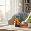 Main tenant un tumbler blanc avec une illustration colorée d un éléphant et de deux petits éléphants, posé sur une table claire à côté d une petite tasse fumante et d une plante, dans un intérieur lumineux