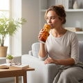 Personne assise dans un fauteuil tenant un tumbler blanc avec une illustration de paysage desertique orange, a cote d une table basse avec un livre ouvert, des lunettes et une petite plante